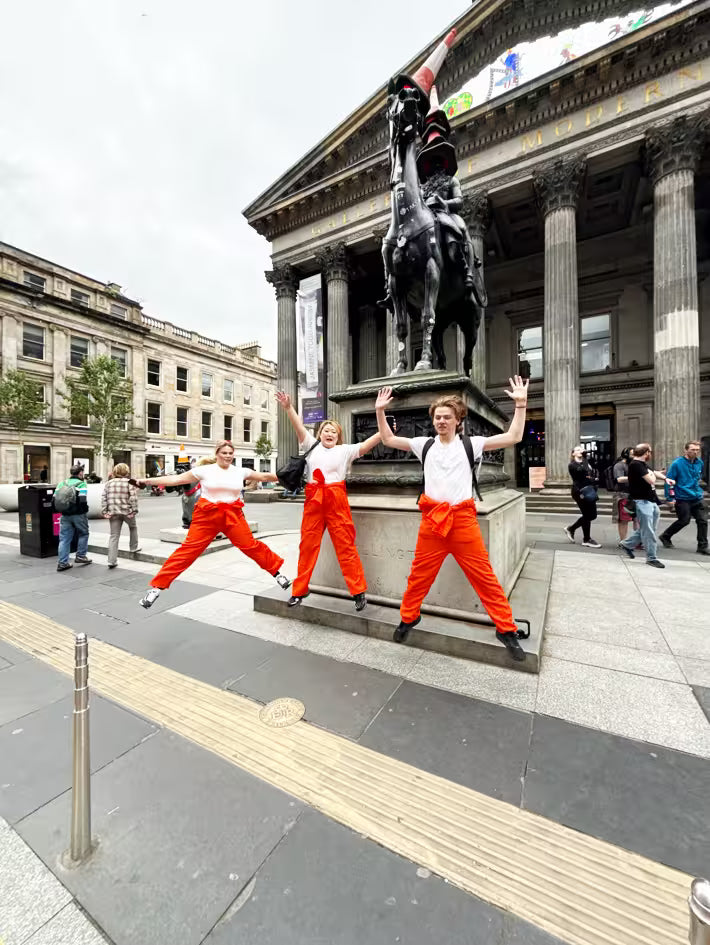 Friends in orange jumpsuits jumping in front of the Duke of Wellington statue in Glasgow on a party treasure hunt