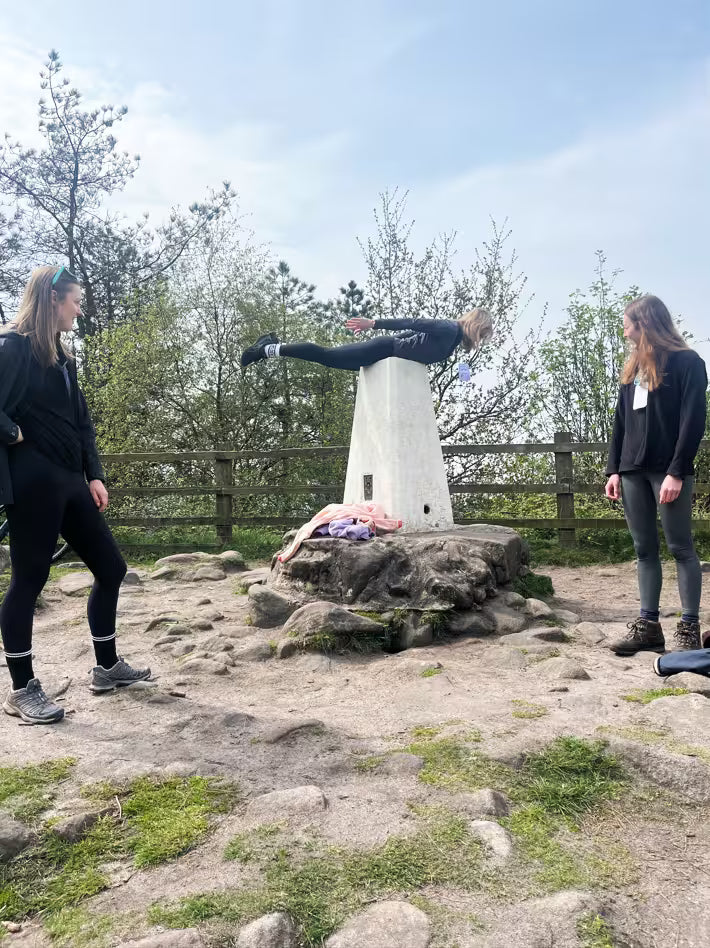Woman planking on top of a hilltop trig point for a treasure hunt photo challenge while her friends watch